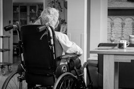 An older person with white hair is sitting in a wheelchair facing away in a cozy indoor setting. A small wooden table nearby holds a glass mug of tea placed on a coaster. The background features some home furnishings and decorative elements.