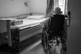 A person in a wheelchair is positioned beside a hospital bed in a simple, sparsely furnished room with a window partially covered by curtains. The room appears to be in a medical or care facility.