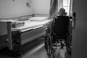 A person in a wheelchair is positioned beside a hospital bed in a simple, sparsely furnished room with a window partially covered by curtains. The room appears to be in a medical or care facility.