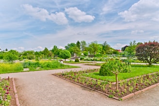 A beautifully maintained garden featuring neatly organized flower beds and lush greenery. Pathways divide the garden into sections, with colorful flowers and well-trimmed bushes adding to the picturesque landscape. The sky above is partly cloudy, providing a serene backdrop.