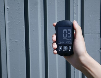 A hand is holding a digital device displaying a reading of 0.3 mmol/L labeled as ketone. The device has a small screen with buttons below it, against a textured, gray, vertical panel background.