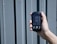 A hand is holding a digital device displaying a reading of 0.3 mmol/L labeled as ketone. The device has a small screen with buttons below it, against a textured, gray, vertical panel background.