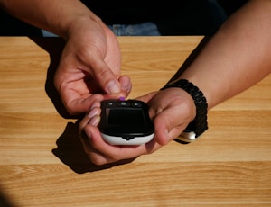 A person is using a glucometer to check blood sugar levels, holding the device with one hand while pricking a finger with a lancet held in the other hand. The hands are positioned over a light wooden surface, and the person is wearing a watch on one wrist.