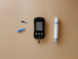 A blood glucose meter is centrally placed on a beige surface, displaying a reading of 103 mg/dL. To its left is a blue lancet for blood sampling, and a black test strip. On the right side is a white and gray lancing device.