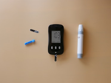 A blood glucose meter is centrally placed on a beige surface, displaying a reading of 103 mg/dL. To its left is a blue lancet for blood sampling, and a black test strip. On the right side is a white and gray lancing device.
