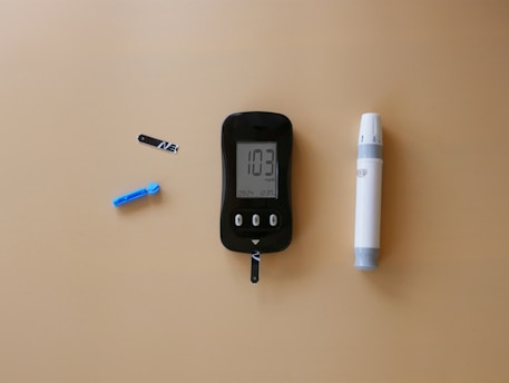 A blood glucose meter is centrally placed on a beige surface, displaying a reading of 103 mg/dL. To its left is a blue lancet for blood sampling, and a black test strip. On the right side is a white and gray lancing device.