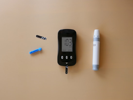 A blood glucose meter is centrally placed on a beige surface, displaying a reading of 103 mg/dL. To its left is a blue lancet for blood sampling, and a black test strip. On the right side is a white and gray lancing device.