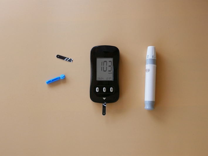 A blood glucose meter is centrally placed on a beige surface, displaying a reading of 103 mg/dL. To its left is a blue lancet for blood sampling, and a black test strip. On the right side is a white and gray lancing device.
