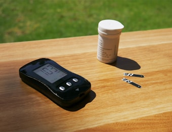 A glucose meter with a digital display reading 103 is placed on a wooden surface. Next to it, there is a small white container and two test strips. Sunlight is casting shadows, creating a relaxed and natural atmosphere.