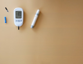 A blood glucose meter displays a reading of 103 mg/dL, accompanied by a lancet and test strip on a plain beige background. The glucometer is white with a digital screen, and there is a cylindrical pen-like device nearby.