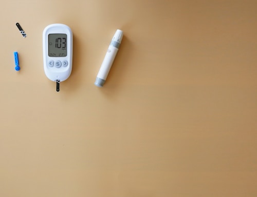A blood glucose meter displays a reading of 103 mg/dL, accompanied by a lancet and test strip on a plain beige background. The glucometer is white with a digital screen, and there is a cylindrical pen-like device nearby.