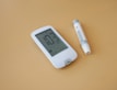 A digital glucose meter and a lancing device are placed on a light brown surface. The glucose meter displays a reading of 103 mg/dL along with other indicators. The lancing device is cylindrical, primarily white with gray accents.
