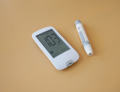 A digital glucose meter and a lancing device are placed on a light brown surface. The glucose meter displays a reading of 103 mg/dL along with other indicators. The lancing device is cylindrical, primarily white with gray accents.