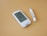 A digital glucose meter and a lancing device are placed on a light brown surface. The glucose meter displays a reading of 103 mg/dL along with other indicators. The lancing device is cylindrical, primarily white with gray accents.