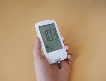 A person holds a white blood glucose meter displaying a reading of 103 mg/dL. The device features a digital screen with additional icons and information.