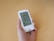A person holds a white blood glucose meter displaying a reading of 103 mg/dL. The device features a digital screen with additional icons and information.