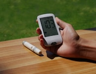 A hand holding a digital glucose meter displaying a reading of 103 mg/dL. The background shows a wooden surface with a pen-like device lying on the table. A grassy lawn is visible in the distance.