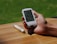 A hand holding a digital glucose meter displaying a reading of 103 mg/dL. The background shows a wooden surface with a pen-like device lying on the table. A grassy lawn is visible in the distance.