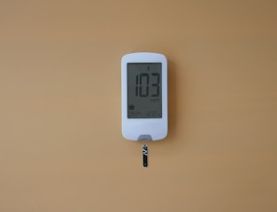 A digital glucose meter displays the reading 103 mg/dL. The device is white with a clear screen and features a test strip inserted at the bottom. The background is a solid mustard yellow color.