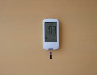 A digital glucose meter displays the reading 103 mg/dL. The device is white with a clear screen and features a test strip inserted at the bottom. The background is a solid mustard yellow color.