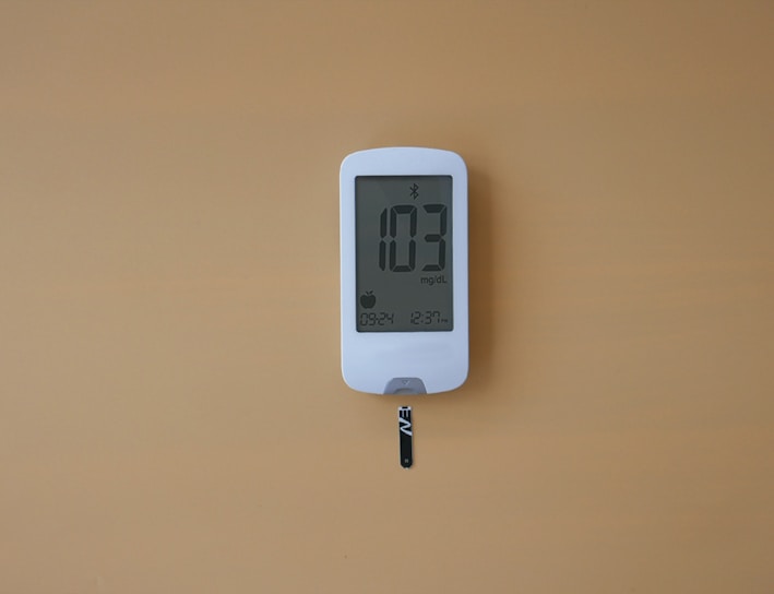 A digital glucose meter displays the reading 103 mg/dL. The device is white with a clear screen and features a test strip inserted at the bottom. The background is a solid mustard yellow color.