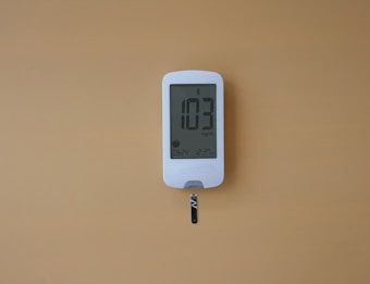 A digital glucose meter displays the reading 103 mg/dL. The device is white with a clear screen and features a test strip inserted at the bottom. The background is a solid mustard yellow color.