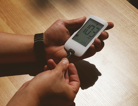 A person's hands are holding a glucose meter displaying a reading of 103 mg/dL. The device is white with a clear digital display. It appears to be on a wooden surface, and there is a test strip being inserted into the meter.