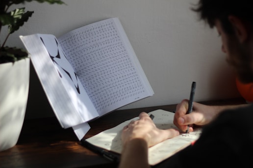 A person is seated at a table writing in a notebook with a pen. An open book filled with various characters or symbols stands upright on the table, possibly being referenced. A potted plant is partially visible in the upper left corner. The scene is well-lit with natural light.