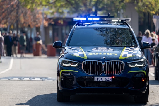A police car with flashing blue lights is positioned on a street. The vehicle has distinctive checkered markings and is a BMW model. In the background, there are several people standing on the sidewalk, blurred, suggesting the photo was taken during daylight in an urban setting.