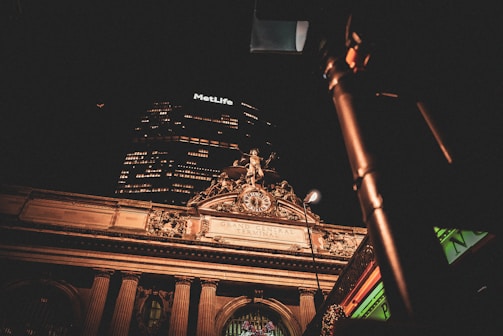 A nighttime view of an illuminated historic building facade with classical architectural details, featuring a clock and sculptures. Above the structure, a modern skyscraper displays the glowing MetLife logo. The scene is partially obscured by an out-of-focus street light in the foreground.