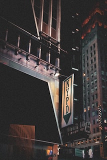 A night scene featuring an urban environment with tall buildings. The NYPD sign is prominently displayed on a building, with some architectural details visible. The signage is illuminated and there are glimpses of other lit-up signs in the background.
