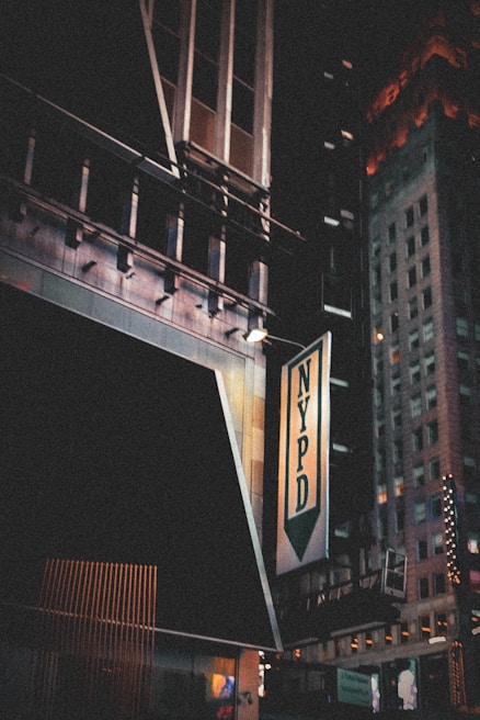 A night scene featuring an urban environment with tall buildings. The NYPD sign is prominently displayed on a building, with some architectural details visible. The signage is illuminated and there are glimpses of other lit-up signs in the background.