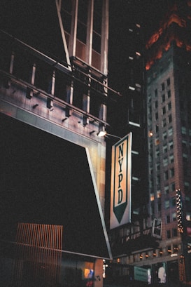 A night scene featuring an urban environment with tall buildings. The NYPD sign is prominently displayed on a building, with some architectural details visible. The signage is illuminated and there are glimpses of other lit-up signs in the background.