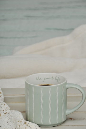 A light green ceramic mug with the phrase 'the good life' written on it sits on a wooden surface, alongside a white knit blanket. The setting appears cozy and comforting, suggesting a relaxing environment.