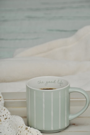 A light green ceramic mug with the phrase 'the good life' written on it sits on a wooden surface, alongside a white knit blanket. The setting appears cozy and comforting, suggesting a relaxing environment.