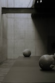 A minimalist interior space is depicted with a concrete wall and floor, featuring large, spherical stone sculptures placed on the floor. The lighting creates dramatic shadows and a calm, quiet atmosphere.