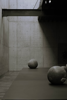 A minimalist interior space is depicted with a concrete wall and floor, featuring large, spherical stone sculptures placed on the floor. The lighting creates dramatic shadows and a calm, quiet atmosphere.