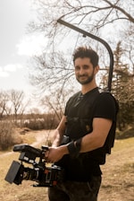 A person outdoors holding a professional camera mounted to a stabilizing harness. Leafless trees in the background and a grassy area suggest an early spring or late autumn setting under a partly cloudy sky.