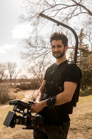 A person outdoors holding a professional camera mounted to a stabilizing harness. Leafless trees in the background and a grassy area suggest an early spring or late autumn setting under a partly cloudy sky.
