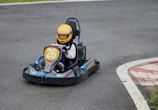 A young child wearing a yellow helmet is driving a small go-kart on an asphalt track. The go-kart has the number 26 on the front and features colorful designs including stars and stripes. The child appears focused on driving as they navigate a curve in the track. The surrounding area includes some grass and a red and white kerb at the edge of the track.