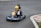 A young child wearing a yellow helmet is driving a small go-kart on an asphalt track. The go-kart has the number 26 on the front and features colorful designs including stars and stripes. The child appears focused on driving as they navigate a curve in the track. The surrounding area includes some grass and a red and white kerb at the edge of the track.