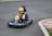 A young child wearing a yellow helmet is driving a small go-kart on an asphalt track. The go-kart has the number 26 on the front and features colorful designs including stars and stripes. The child appears focused on driving as they navigate a curve in the track. The surrounding area includes some grass and a red and white kerb at the edge of the track.
