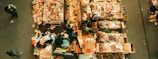 An aerial view of a flea market stall with tables filled with assorted items. Various people are walking around, inspecting goods. The tables display a mix of vintage and second-hand items, including books, clothes, pottery, and knick-knacks.
