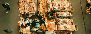 An aerial view of a flea market stall with tables filled with assorted items. Various people are walking around, inspecting goods. The tables display a mix of vintage and second-hand items, including books, clothes, pottery, and knick-knacks.