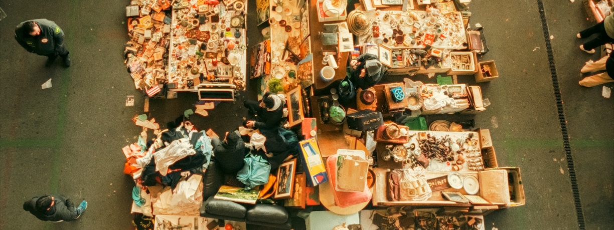 An aerial view of a flea market stall with tables filled with assorted items. Various people are walking around, inspecting goods. The tables display a mix of vintage and second-hand items, including books, clothes, pottery, and knick-knacks.