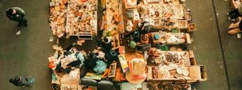 An aerial view of a flea market stall with tables filled with assorted items. Various people are walking around, inspecting goods. The tables display a mix of vintage and second-hand items, including books, clothes, pottery, and knick-knacks.