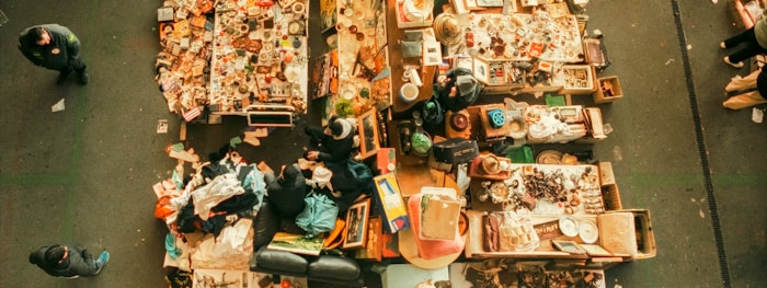 An aerial view of a flea market stall with tables filled with assorted items. Various people are walking around, inspecting goods. The tables display a mix of vintage and second-hand items, including books, clothes, pottery, and knick-knacks.