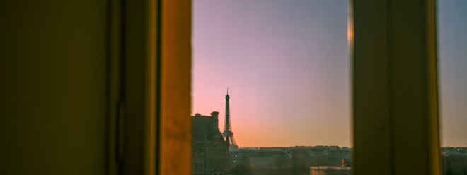 A view through a window frame captures a cityscape at sunset. The Eiffel Tower is prominently visible against a gradient sky transitioning from soft pink to orange. The outlines of surrounding buildings are silhouetted in the foreground.