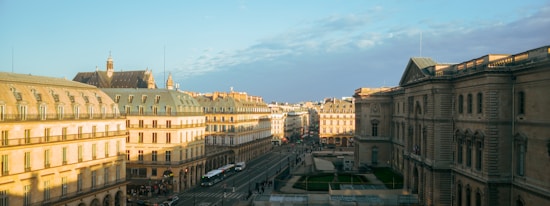 A cityscape featuring historic European architecture with buildings lining both sides of a central street. The sunlight casts a warm glow on the facades, highlighting intricate architectural details such as windows, roofs, and chimneys. There are people walking and cars driving on the street, adding vibrant life to the scene.