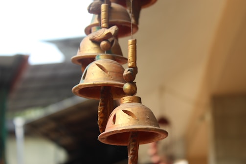 Several metallic bells hang in a vertical line. The bells have a rustic, antique appearance and are set against a blurred background, suggesting they might be a type of wind chime or decorative element.